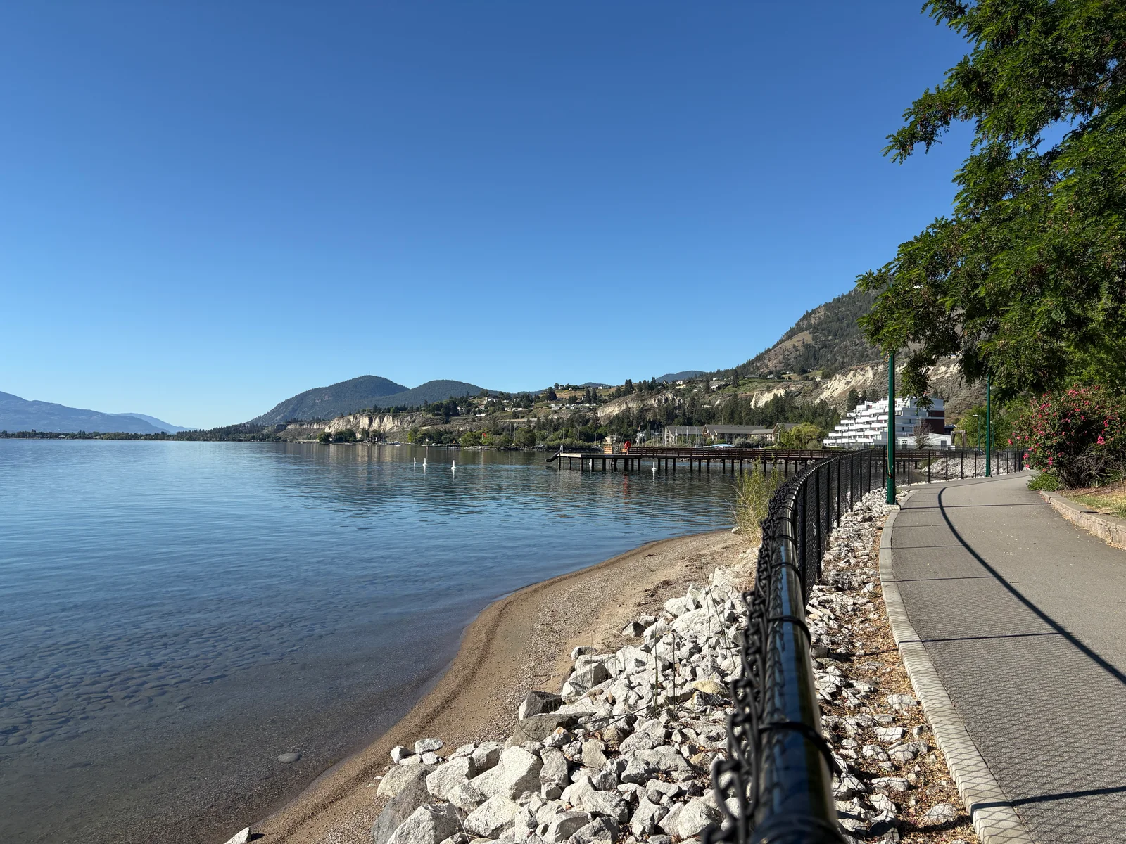 Okanagan waterfront with calm blue water, hillside cliffs, and a lakeside walking path